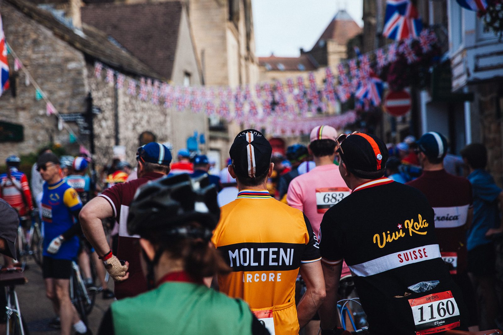 Cyclists gather in a quaint, decorated village, lined with colorful bunting and flags. Riders wear vintage-style jerseys and helmets, with visible bib numbers. The scene captures a festive, communal atmosphere.
