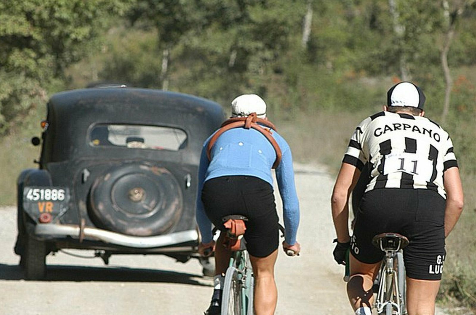 Two cyclists in vintage attire ride on a dirt road behind an old black car with the license plate "451886 VR." One cyclist wears a blue long-sleeve shirt, while the other wears a white and black striped jersey with "CARPANO" and "11" written on it. Trees line the road ahead.