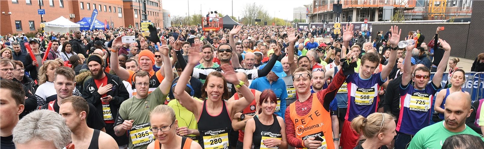 A large group of runners, wearing race bibs and athletic gear, smile and wave at the camera as they prepare to start a race. Buildings and spectators are visible in the background. The atmosphere appears energetic and enthusiastic.