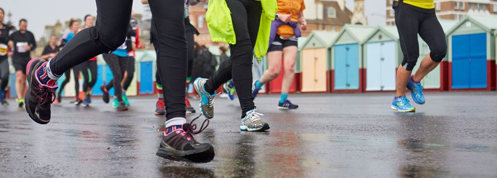 Runners on a wet surface participating in an outdoor event. They are dressed in athletic gear, such as leggings and sneakers. Colorful beach huts line the background, hinting at a coastal location. The image shows movement and energy.