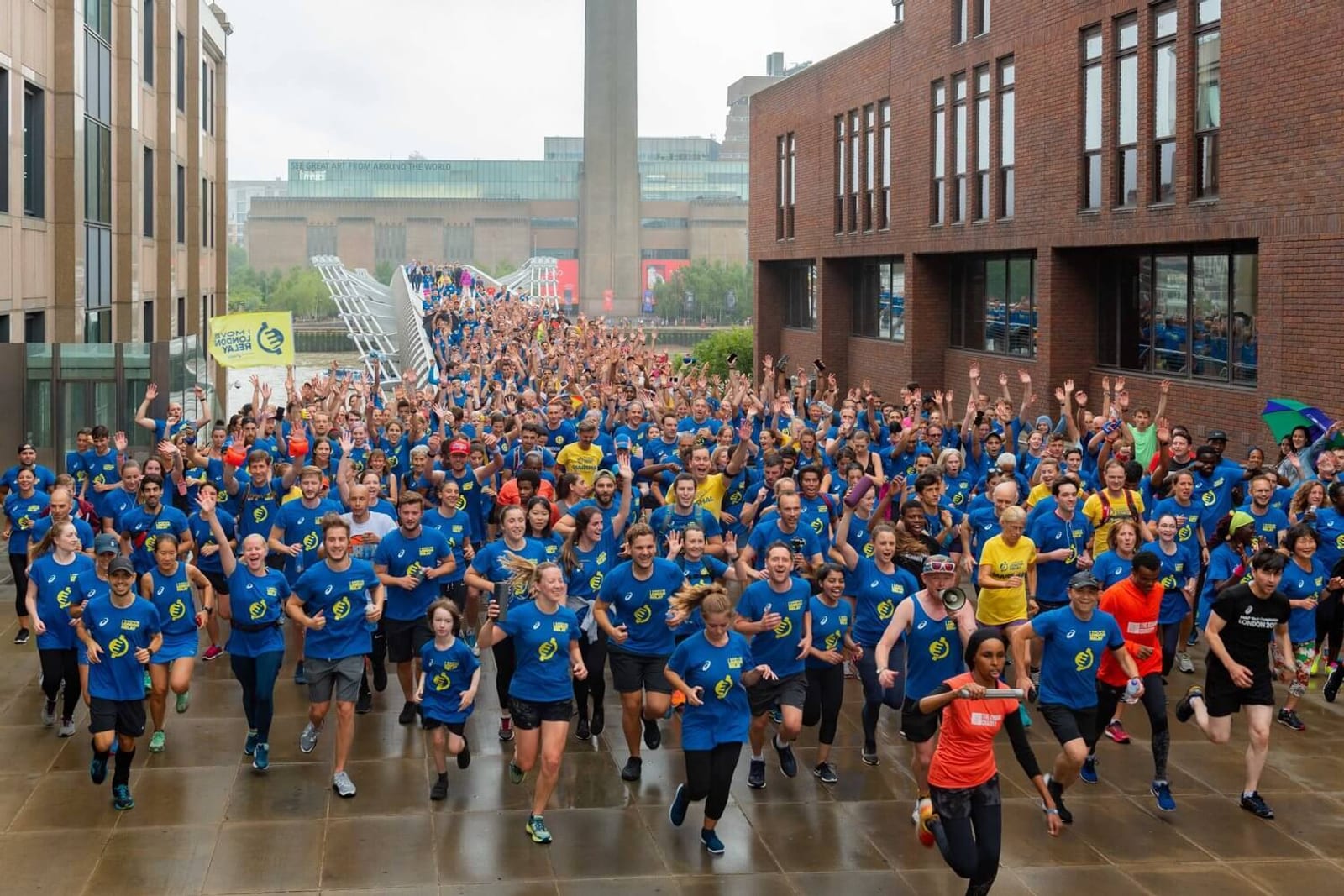 Hundreds of people in blue shirts participate in a large outdoor fun run, smiling and cheering as they run on a wet pavement between modern buildings on a cloudy day.