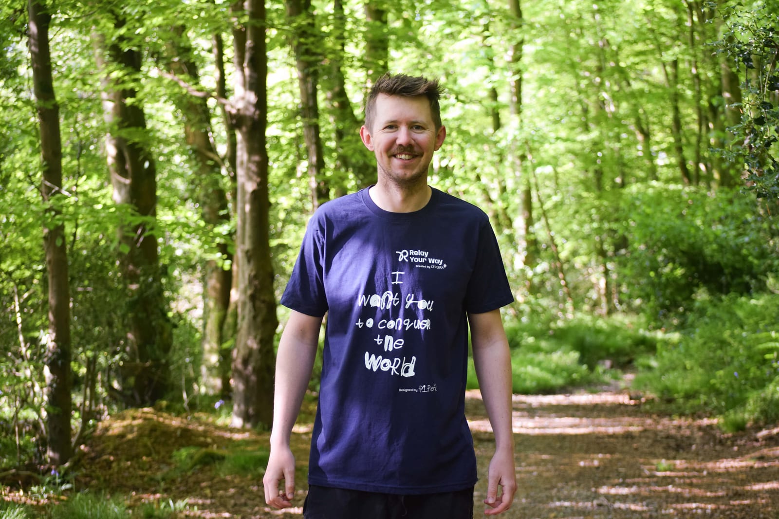 A man with short hair and a mustache smiles while standing on a sunlit forest path, wearing a navy blue t-shirt with white text. Green trees and foliage surround the trail in the background.