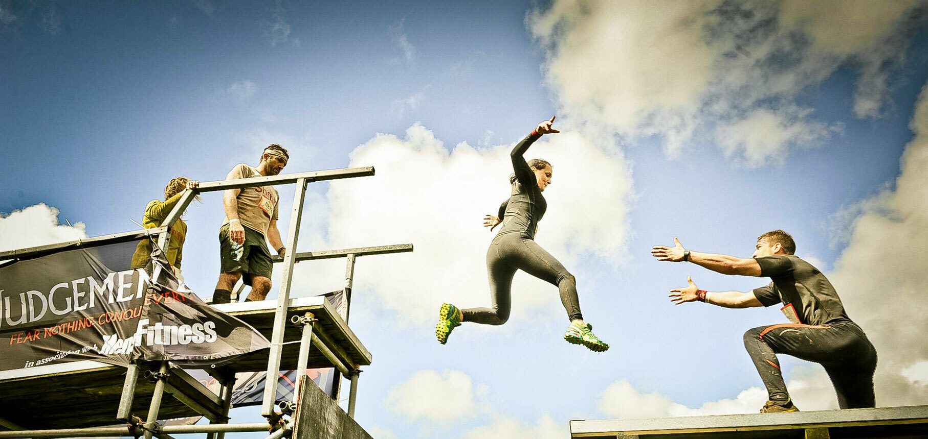 A woman in sportswear leaps across a gap between two elevated platforms, reaching for a man's outstretched hands on the other side. Two people stand on the left platform next to a banner that reads "JUDGEMENT: FEAR NOTHING CONQUER ALL." The sky is partly cloudy.