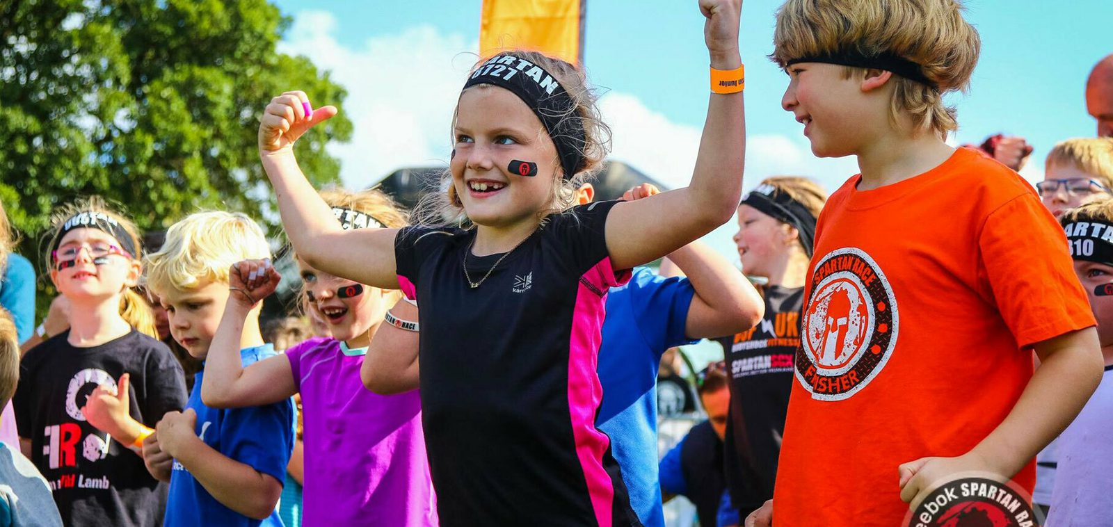 A group of children excitedly participate in an outdoor event. A girl in the center wearing a headband raises her arms in celebration. Other children around her, some also in headbands and event shirts, show expressions of joy and enthusiasm. Trees and a blue sky are in the background.