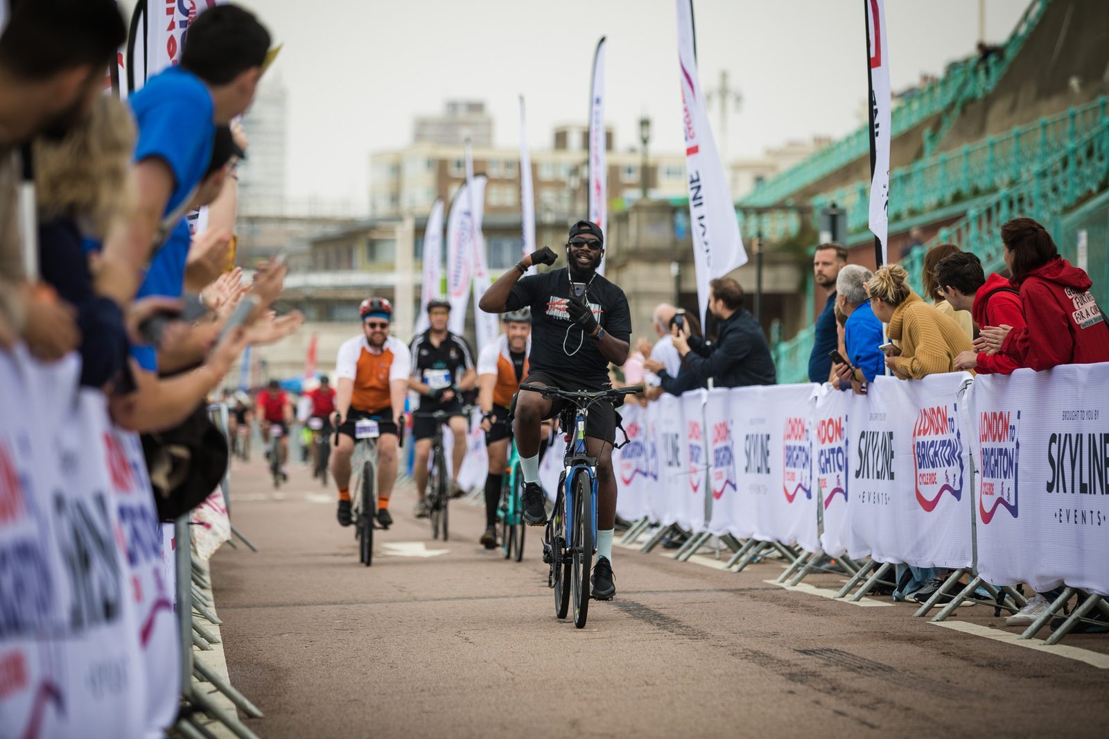 Cyclists participating in a race near a waterfront area, with onlookers cheering them on from the sidelines. The cyclists are riding along a course lined with white banners bearing event logos, and the atmosphere is celebratory.