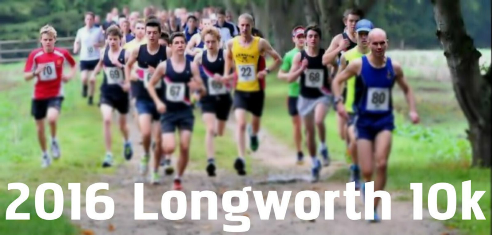 A group of runners is competing in the 2016 Longworth 10k race. The participants, wearing race bibs and various athletic outfits, are running along a tree-lined path. The foreground focuses on the leading runners, with others following closely behind.