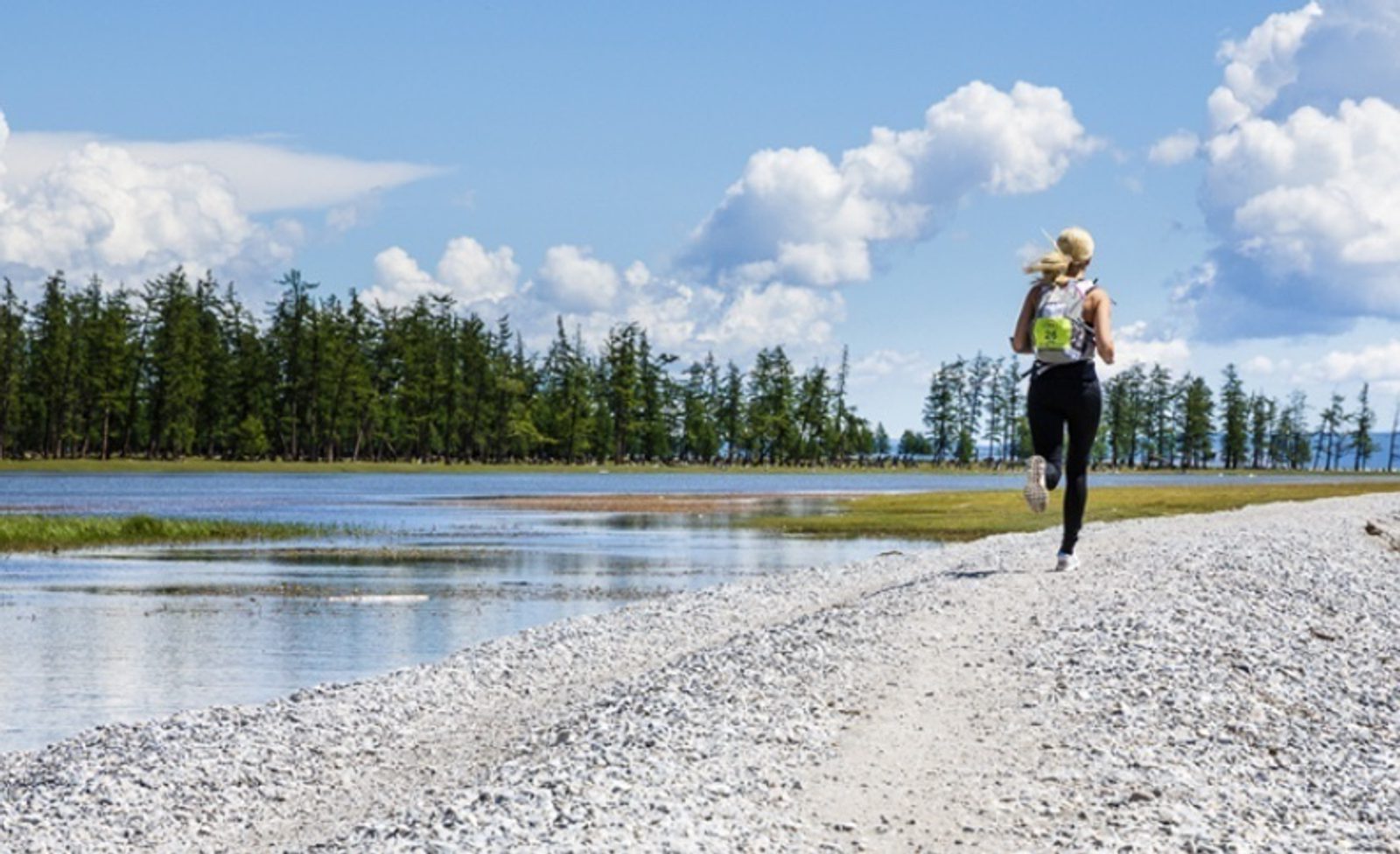 A person with long hair runs on a gravel path alongside a serene body of water, bordered by tall pine trees under a bright blue sky with scattered clouds. They wear athletic clothing and a race bib on their back.