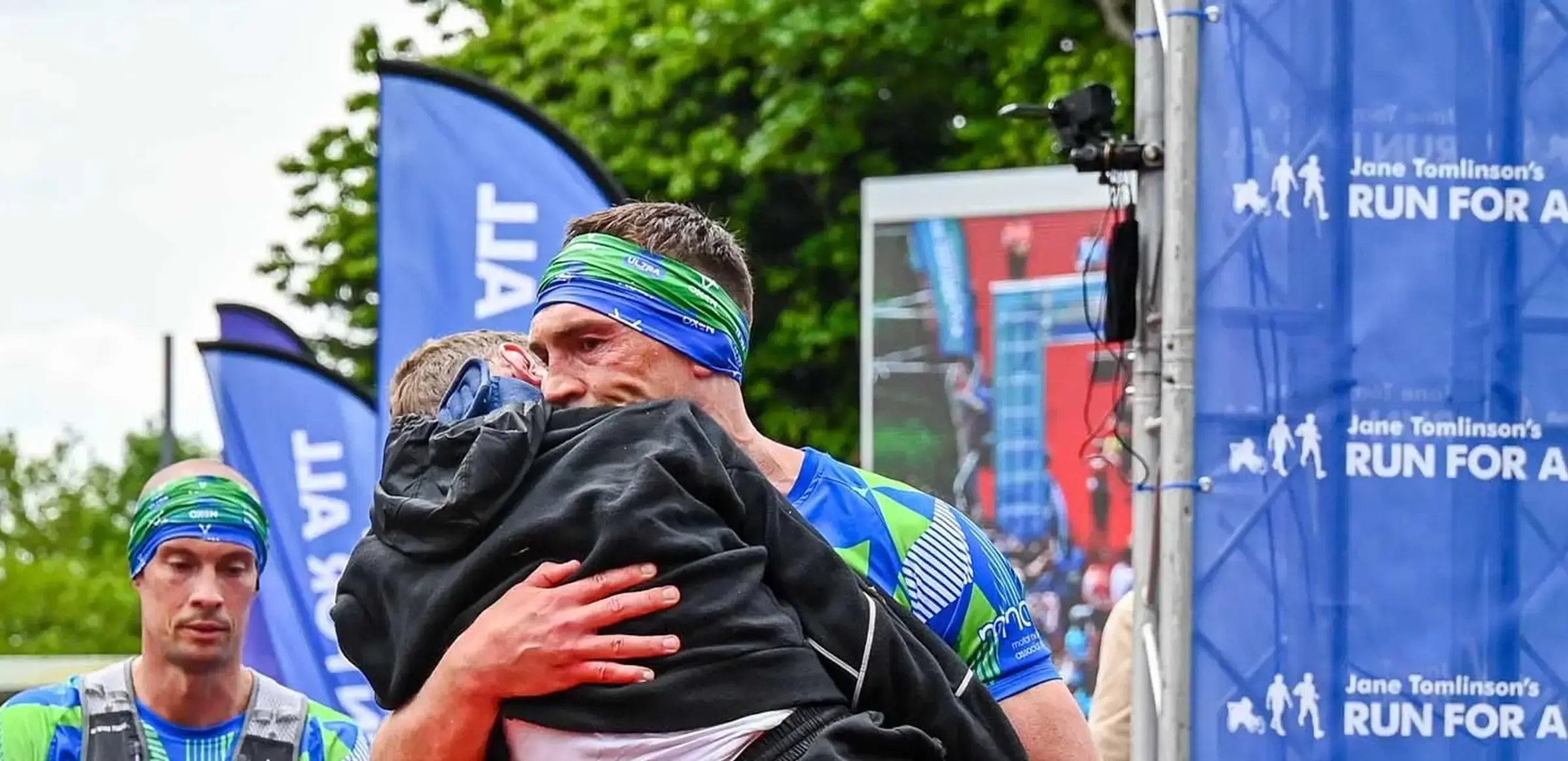 A runner wearing a blue and green headband hugs someone wrapped in a black jacket at a race event. Blue banners and a sign say "Jane Tomlinson's Run For All" in the background, with trees and a large screen visible. Another person with a headband stands nearby.