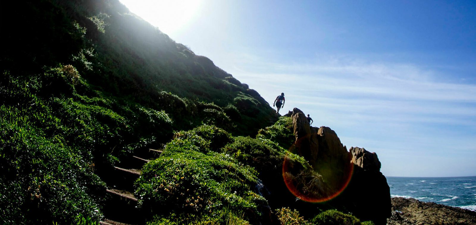 A person stands on a rocky, green hillside under a bright sun with the blue sky and ocean in the background. The hillside features narrow, uneven steps leading upwards. The sun creates a lens flare near the person.