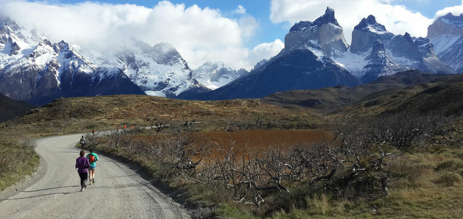 A group of hikers walk along a dirt path winding through mountainous terrain. Snow-capped peaks tower in the background under a partly cloudy sky. Sparse vegetation and a small, murky lake are on either side of the path. The landscape is rugged and scenic.