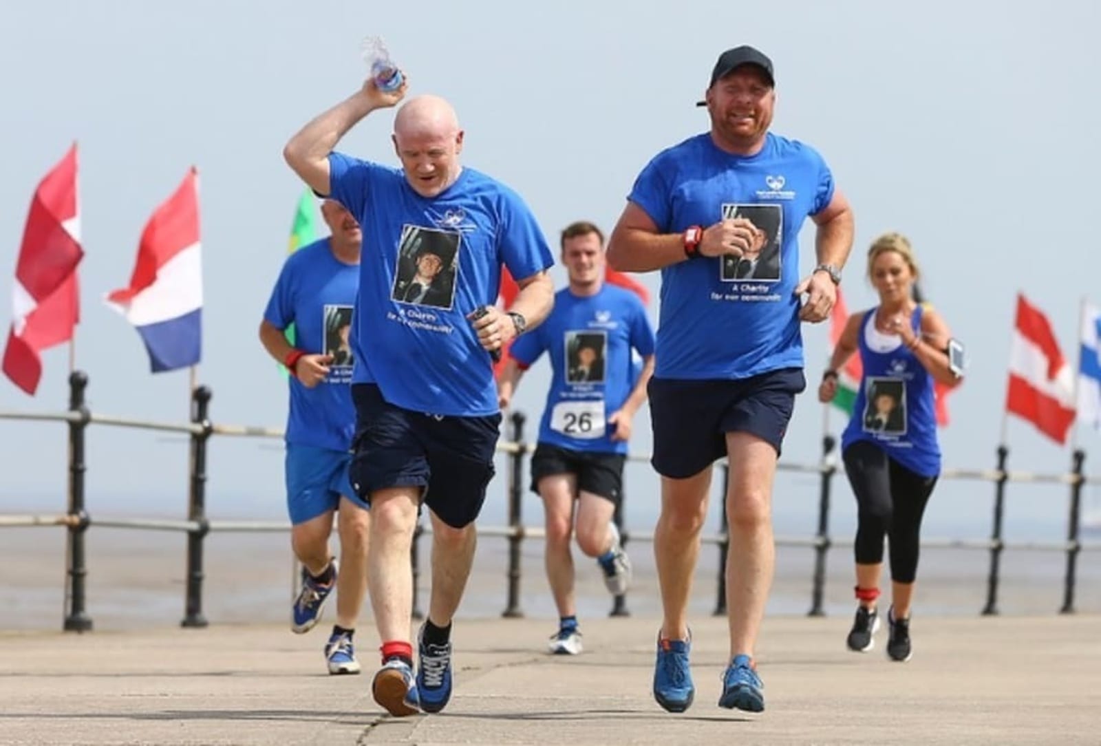 A group of people in blue shirts running on a paved path with various international flags in the background. Two men leading the group are smiling, with one holding a water bottle. They all wear bib numbers and a printed photo on their shirts.