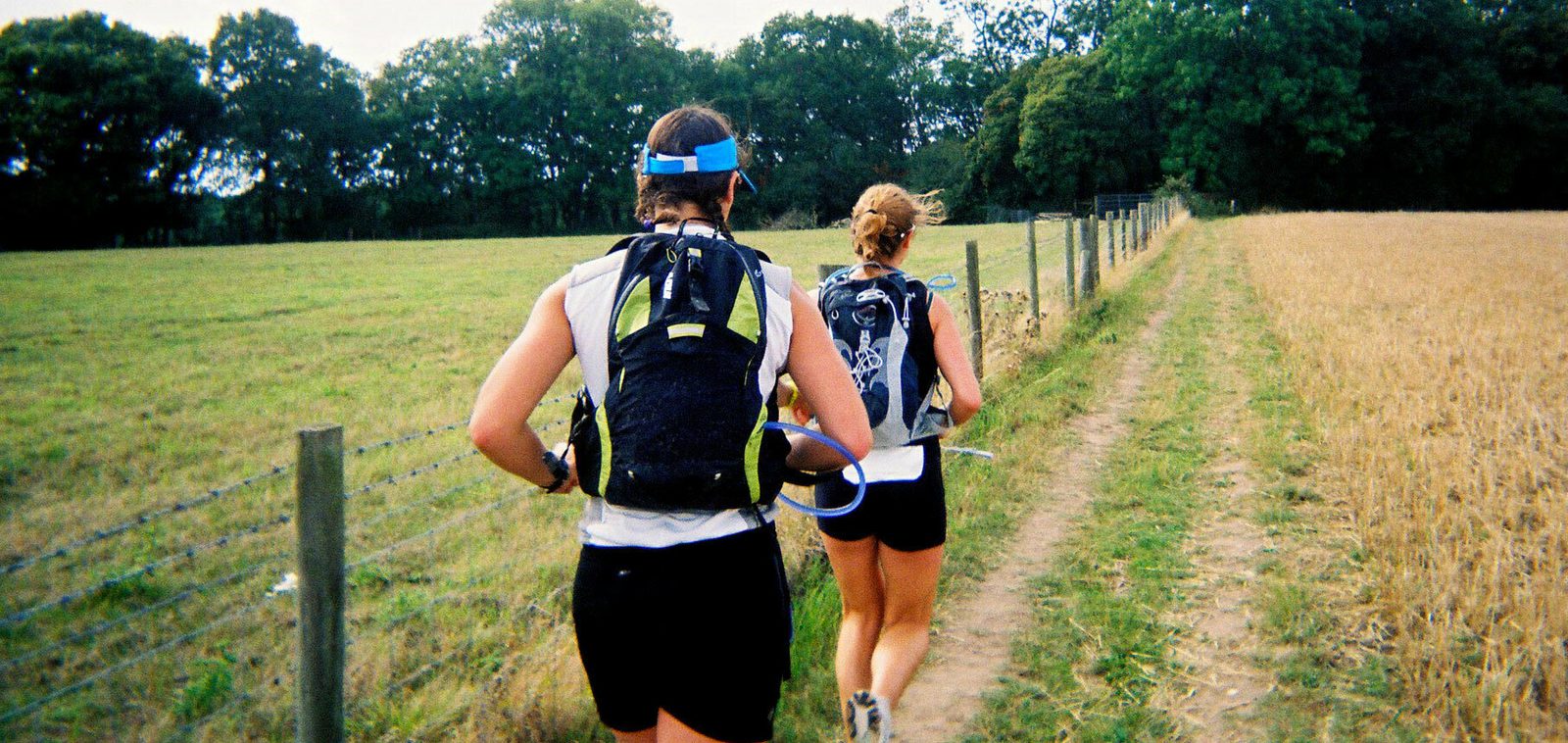 Two runners in athletic gear, including hydration backpacks, run on a dirt trail adjacent to a fence. They are on a path between a grassy field and a harvested one, with trees in the background and a partly cloudy sky overhead.