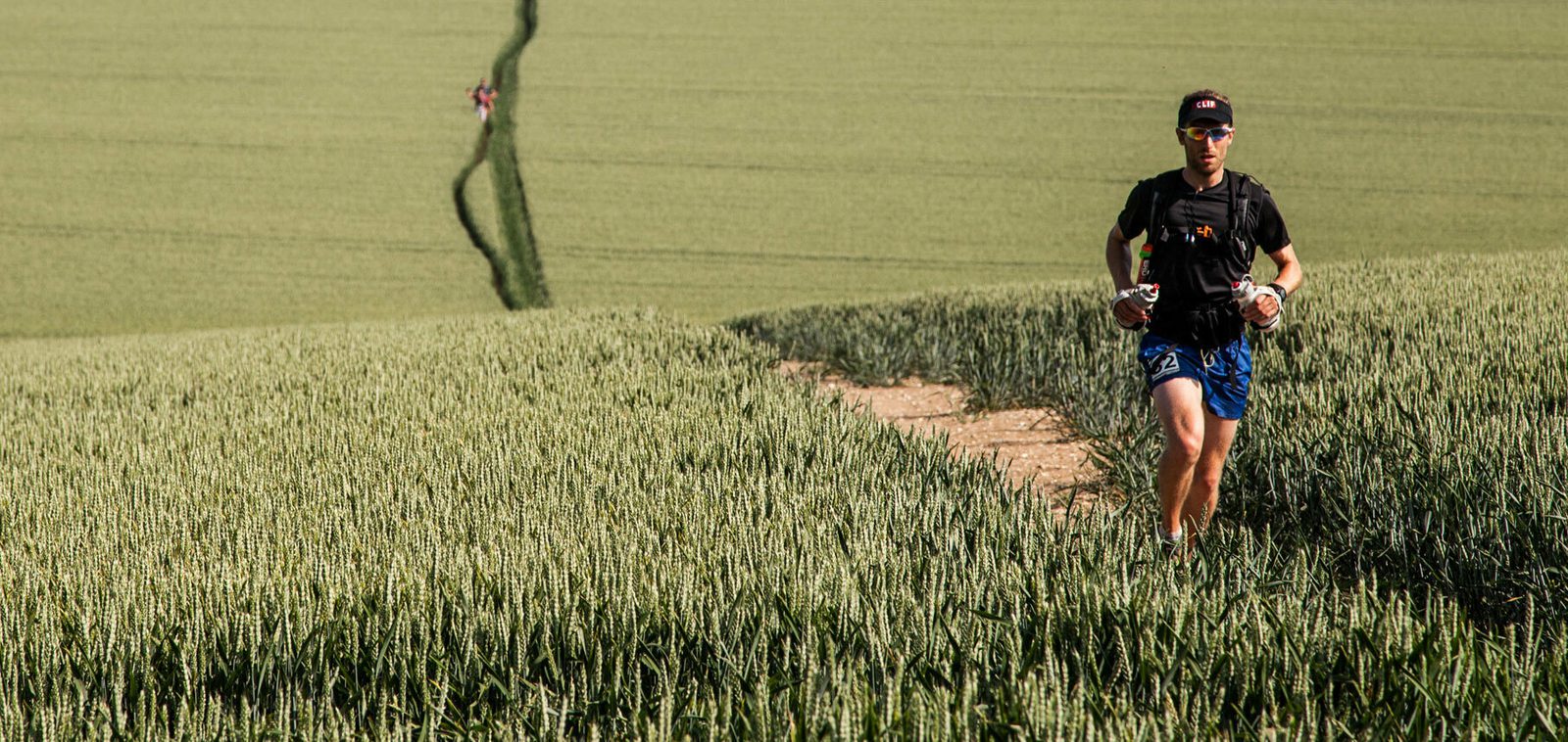 A person in athletic gear runs on a dirt path through a vast green field. Another runner is visible in the background, following the same path. The scene is bright and the sky is clear. Both runners wear gear appropriate for long-distance running or a trail race.