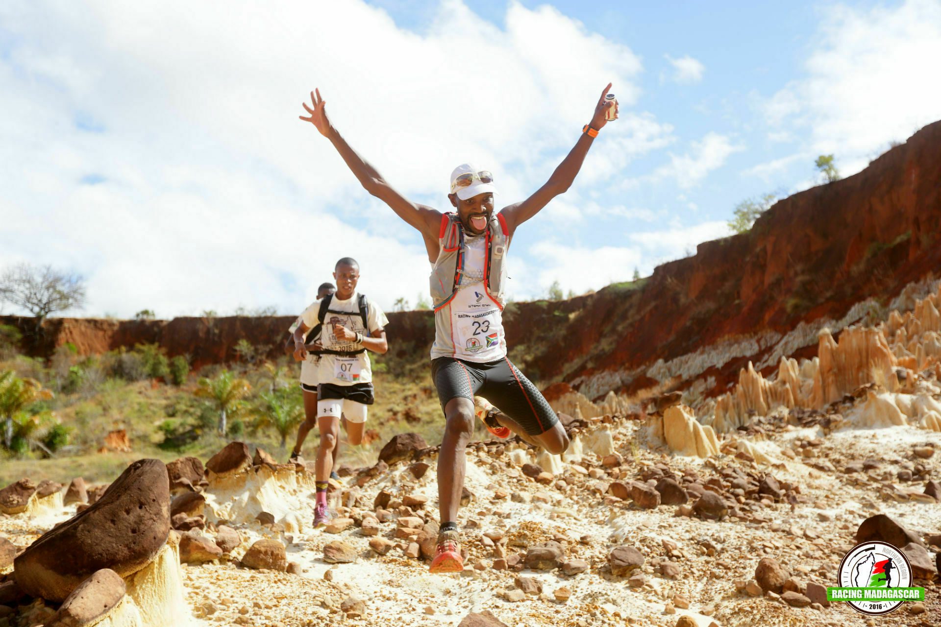 Two athletes race through a rocky, desert-like terrain with arms raised in triumph. The lead runner is airborne, leaping over rough ground. They wear numbered race bibs and vests. The backdrop features rugged cliffs and scattered greenery, with a bright, partly cloudy sky.