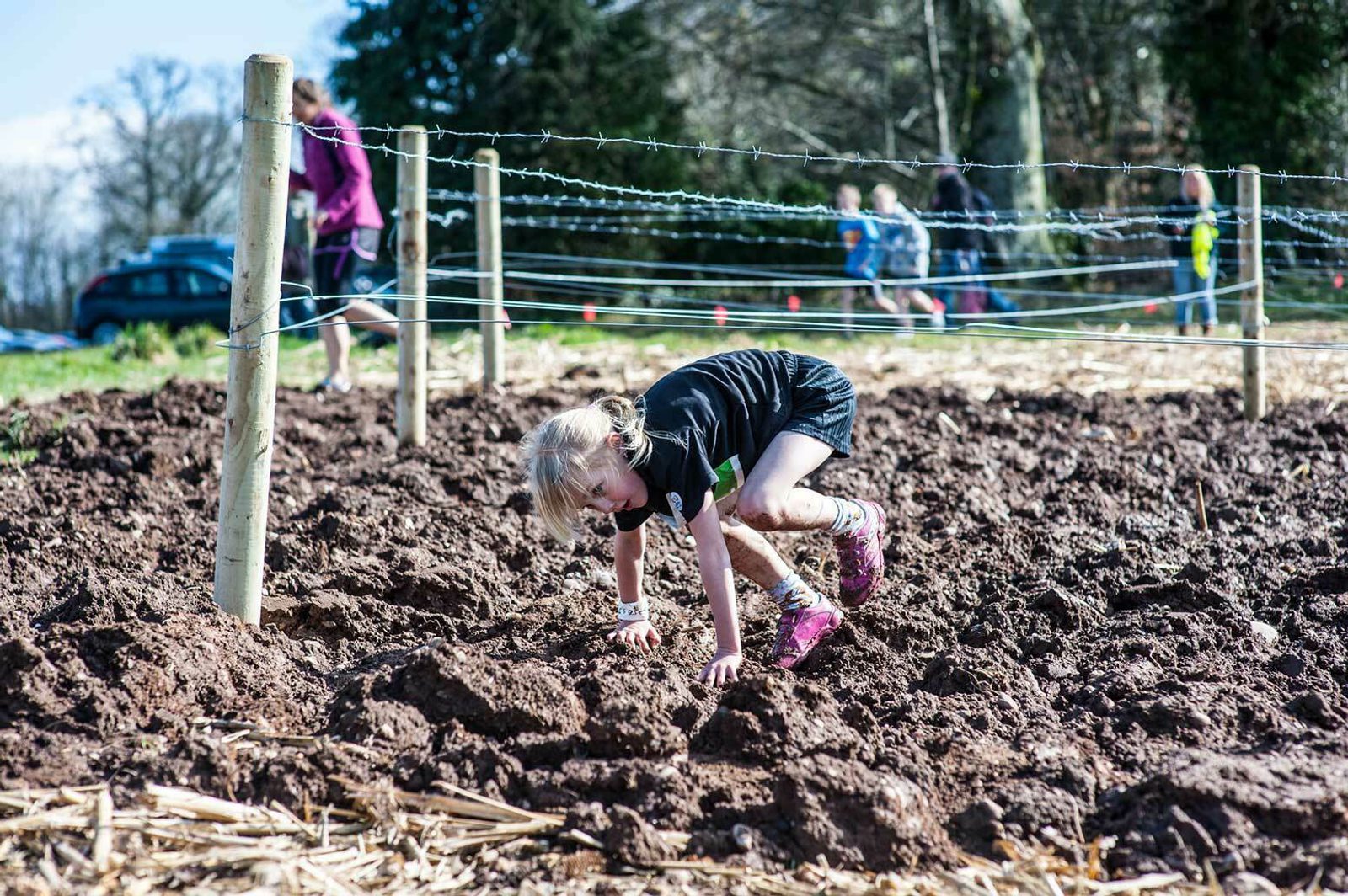 A young child wearing a black shirt and pink shoes is crawling through a muddy obstacle course. Three other people can be seen in the background, navigating the course and passing by a section with barbed wire. Trees and foliage are visible in the distance.