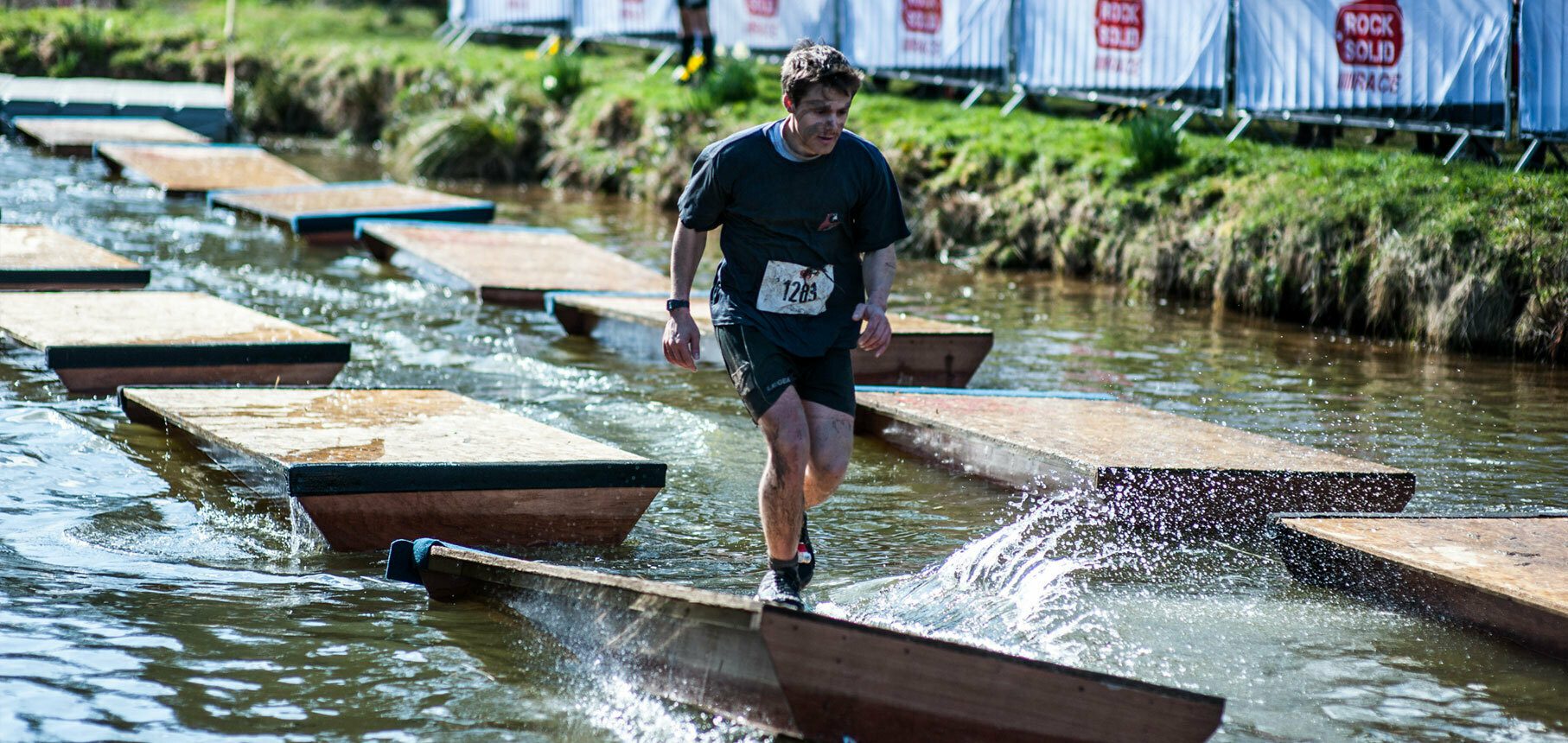 A man wearing a race bib labeled "1264" is crossing a water obstacle during an outdoor race event. He is balancing on a narrow wooden beam with platforms on either side, splashing water as he steps. Spectators and race banners can be seen in the background.