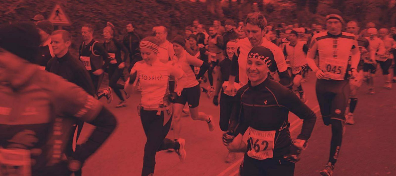 A large group of runners participate in a race on a road lined with trees. Many runners wear numbered bibs, athletic gear, and a variety of hats and headbands. The scene is busy with movement and energy. Some spectators can be seen in the background.