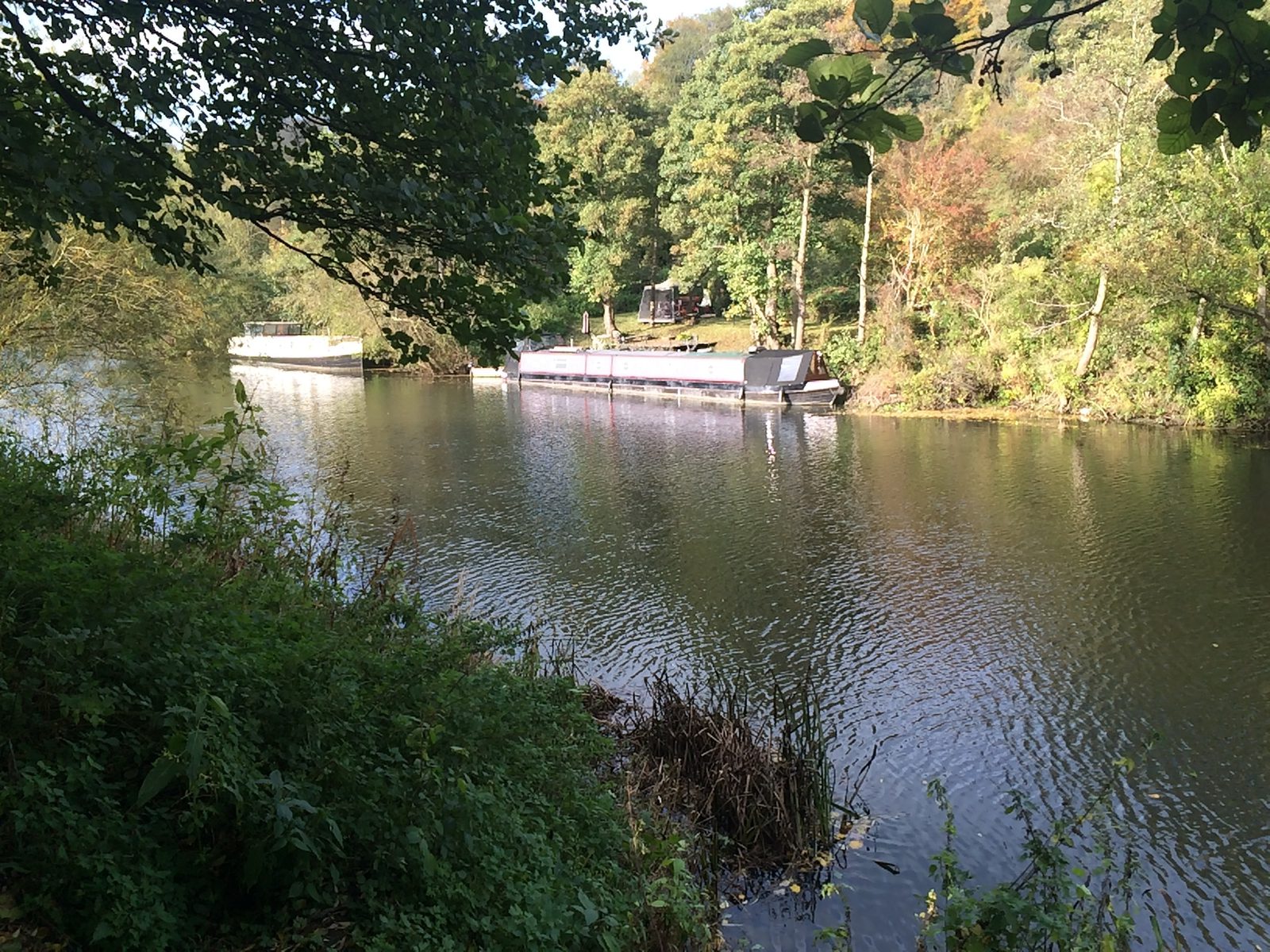 A calm river is flanked by lush greenery and trees with a barge and a smaller boat moored along the bank. The sunny day casts reflections of the boats and foliage on the water's surface, creating a serene and picturesque scene.