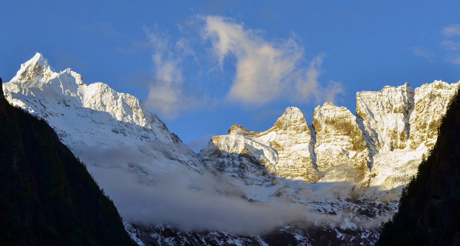 A scenic view of snow-capped mountains under a clear blue sky. The peaks glisten in the sunlight, contrasting sharply with the dark, forested slopes in the foreground. Wisps of clouds drift near the summits, adding to the majestic atmosphere.
