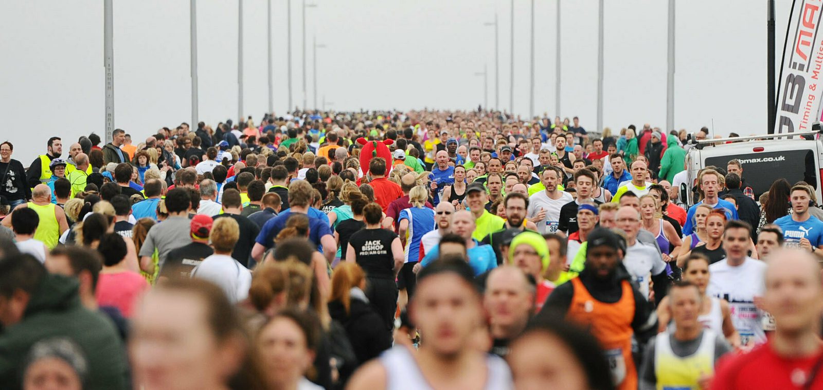 A large crowd of runners participating in a marathon on a foggy day. The runners wear various bright-colored athletic clothing and some costumes. The road is lined with light poles, and the background is filled with more participants running towards the viewer.