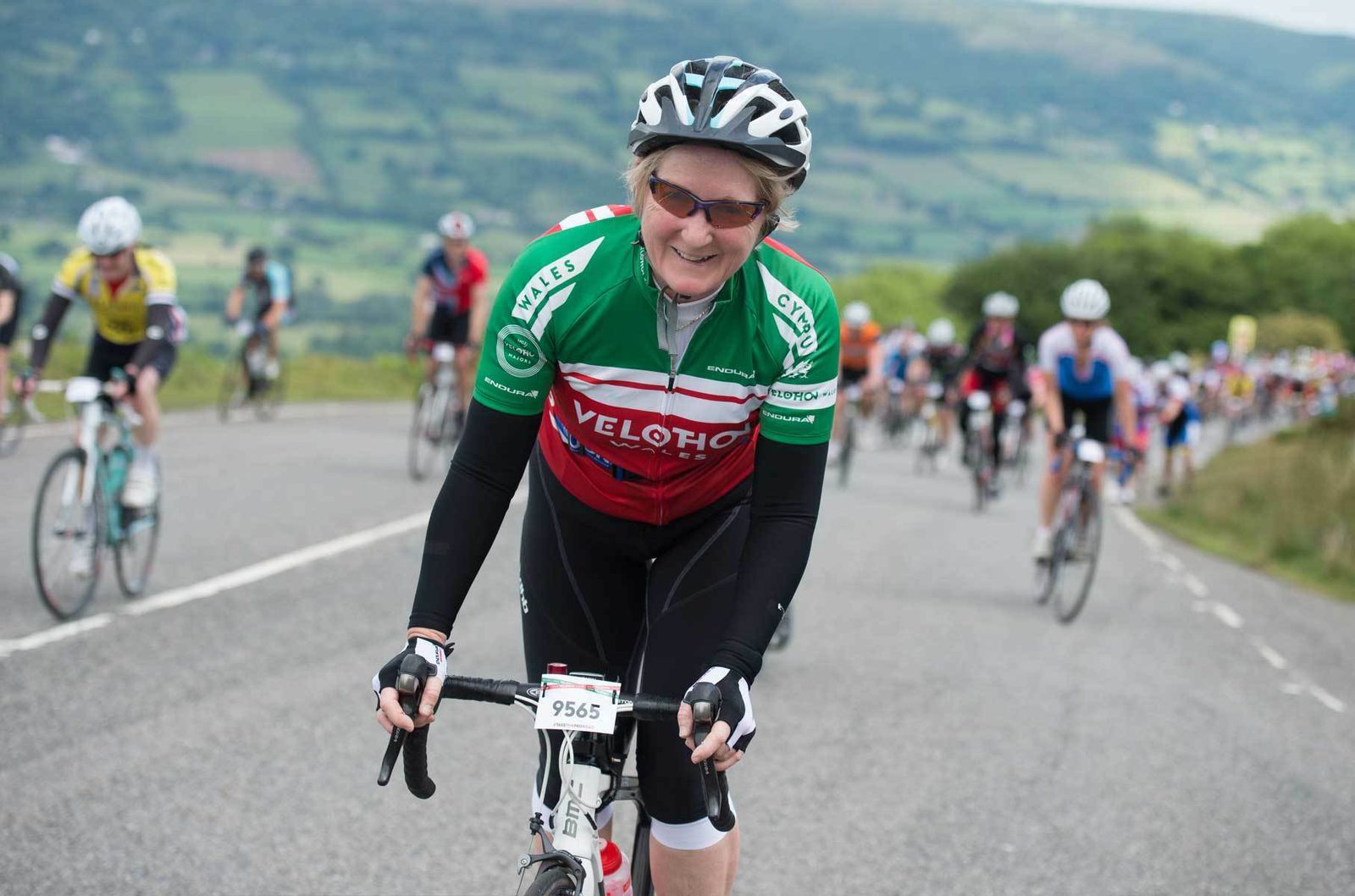 A cyclist in a red, green, and white jersey rides up a hill during a race. The cyclist, wearing sunglasses and a helmet, smiles at the camera. Other cyclists can be seen in the background on the winding road with a scenic, hilly landscape.