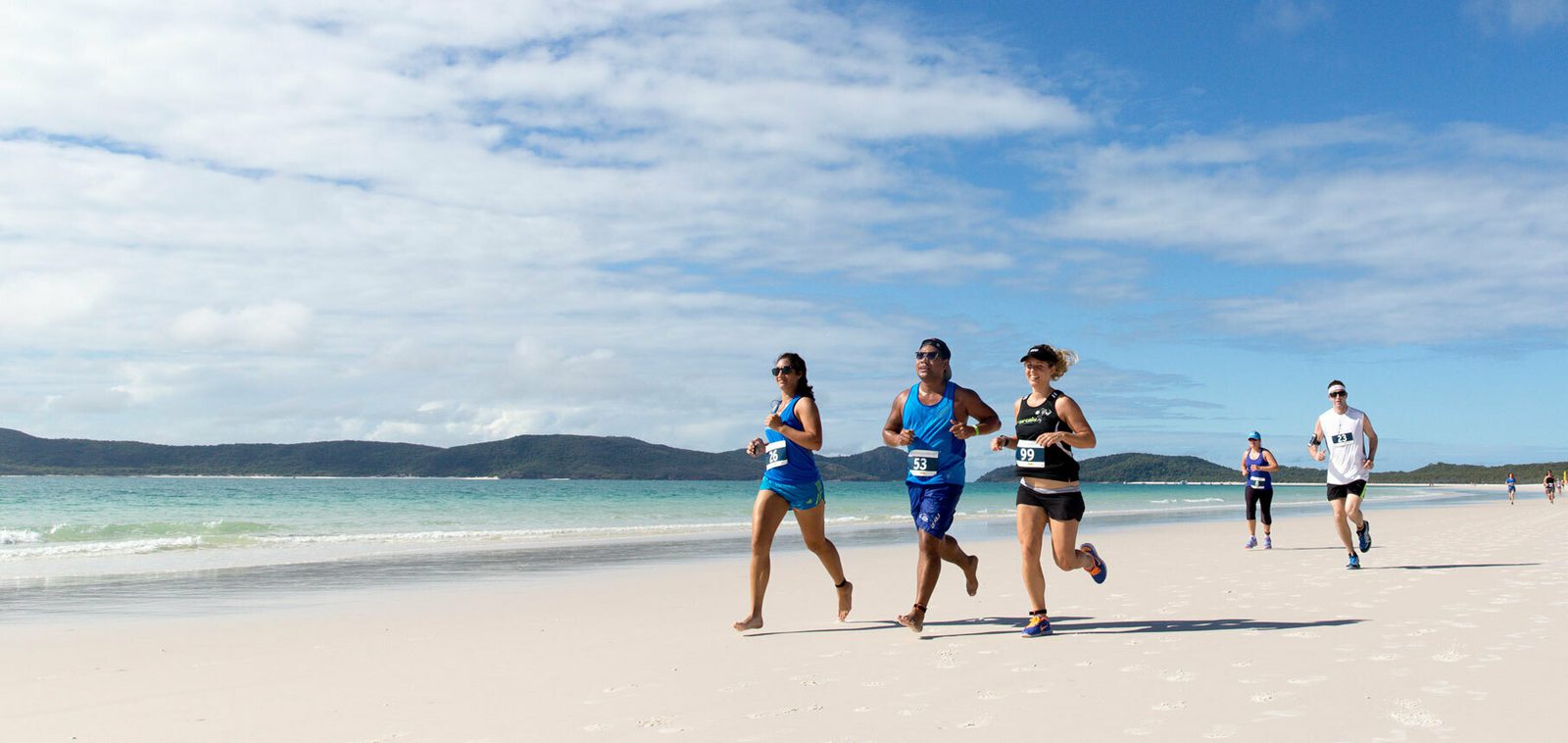 Four people are running along a sandy beach with a clear ocean and distant hills in the background. They are wearing athletic clothing and numbered race bibs, participating in an organized run under a partly cloudy sky.