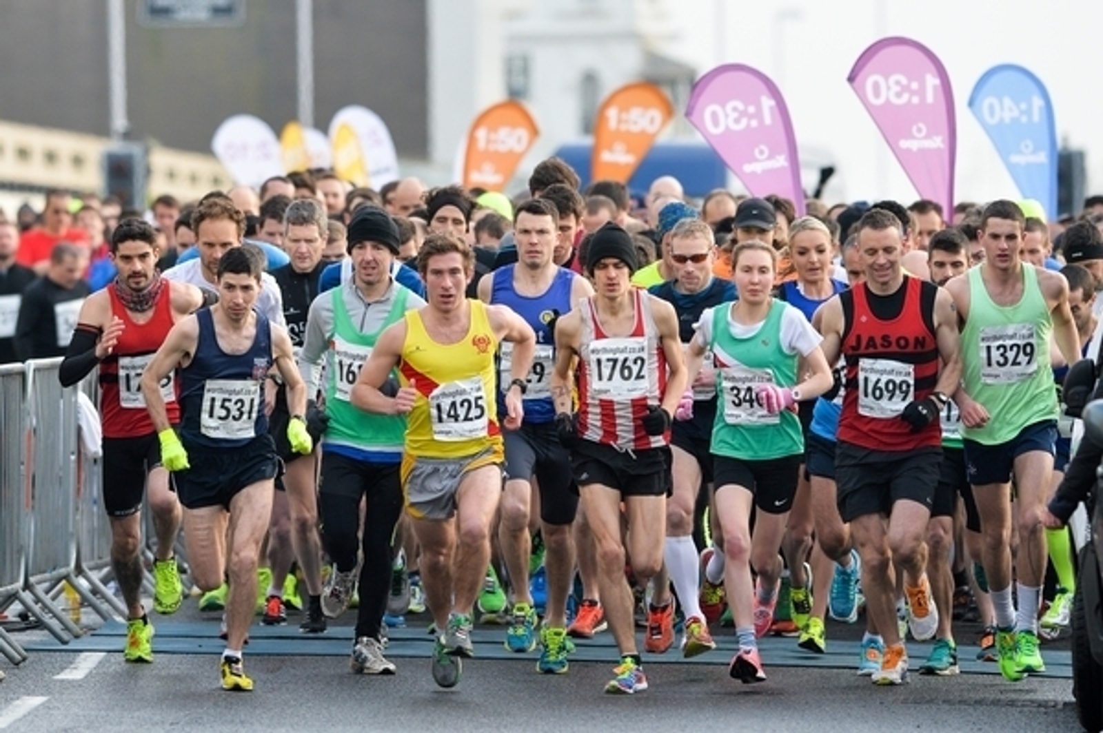 A large group of runners, wearing race bibs and athletic gear, are starting a marathon. Colorful pace marker flags with different times are visible in the background. The race appears to be taking place in an urban area early in the day.