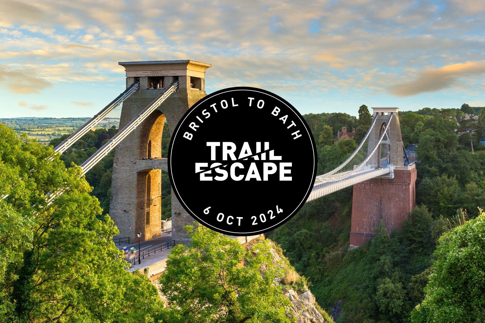 A scenic view of the Clifton Suspension Bridge against a backdrop of a partly cloudy sky. In the center is a black circular logo with text: "Bristol to Bath Trail Escape, 6 Oct 2024." The bridge is surrounded by lush green trees.