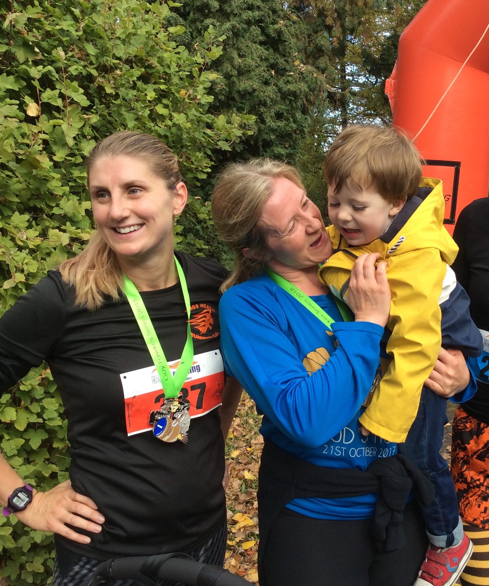 Two women with green medals stand outdoors near greenery. The woman on the left, in a black shirt and race bib, is smiling. The woman on the right, in a blue shirt, is holding a young child in a yellow jacket. An orange structure is visible in the background.
