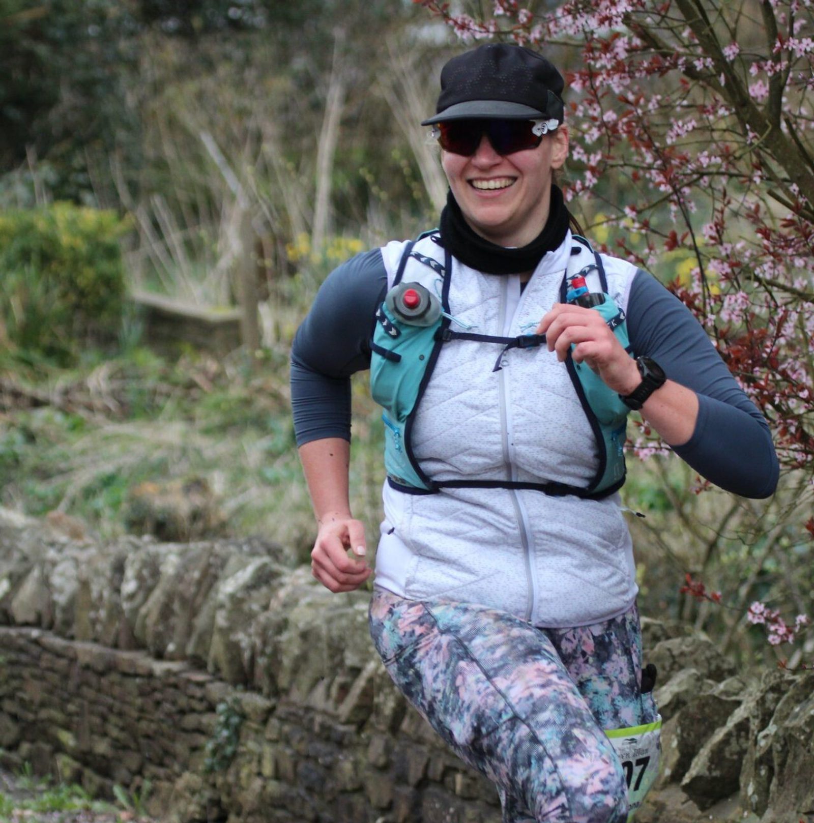 A person is jogging outdoors along a stone wall, wearing a white vest, blue hydration pack, floral-patterned leggings, a black cap, and sunglasses, and smiling while passing by blossoming trees.