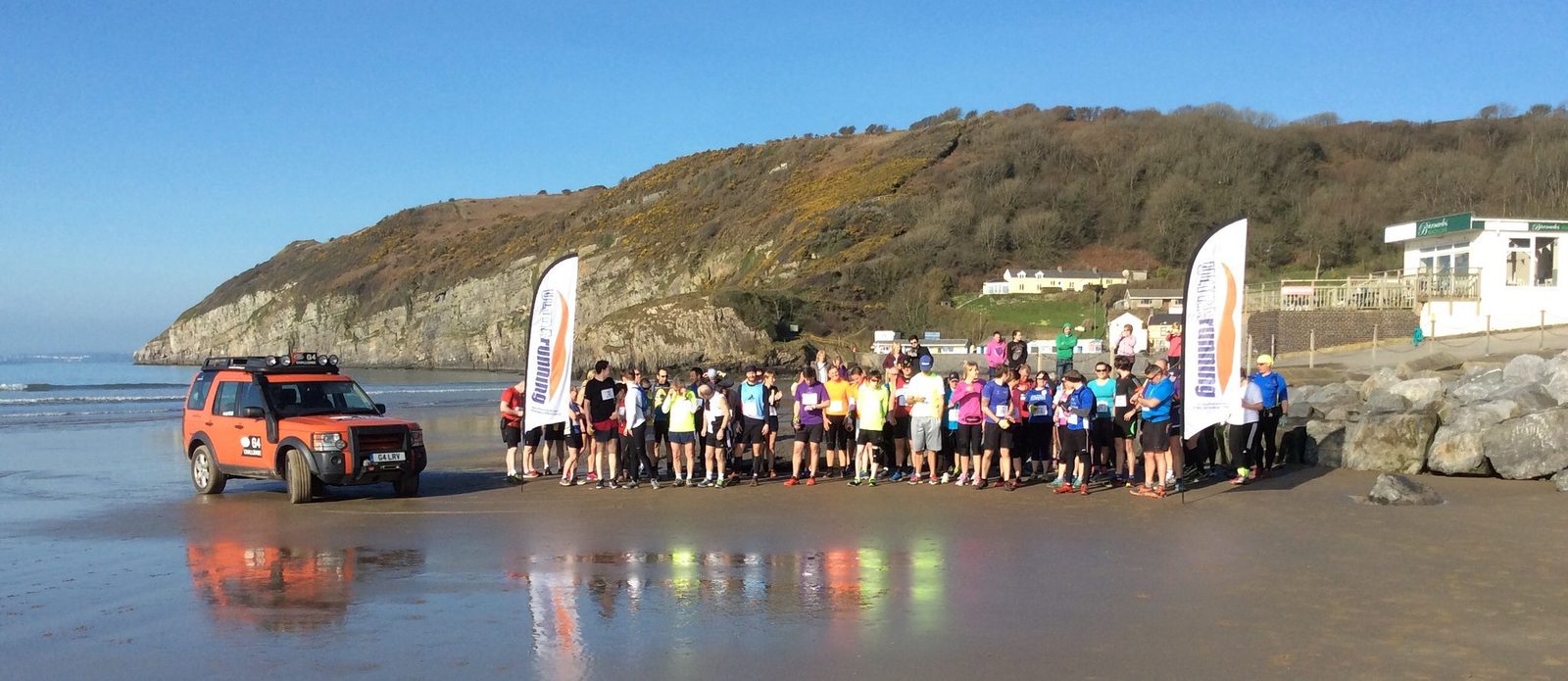 A group of runners is gathered at the start line on a beach, with a safety vehicle parked beside them. They are standing next to two vertical banners. In the background are rocky cliffs and hilly terrain, with a few buildings visible near the shore. The weather is clear and sunny.