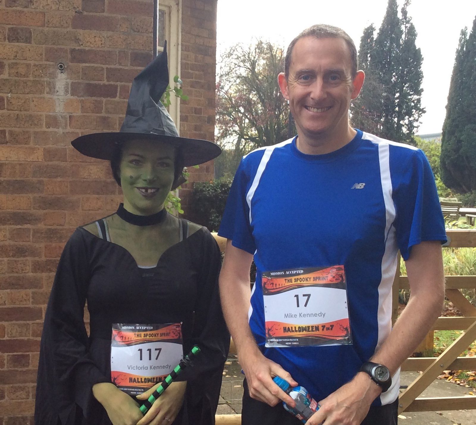 Two people are posing for a photo at a Halloween-themed running event. The person on the left is dressed as a witch with green face paint and a pointed hat, while the person on the right is in athletic wear. Both are holding what appear to be water bottles. They have race bibs pinned to their chests.