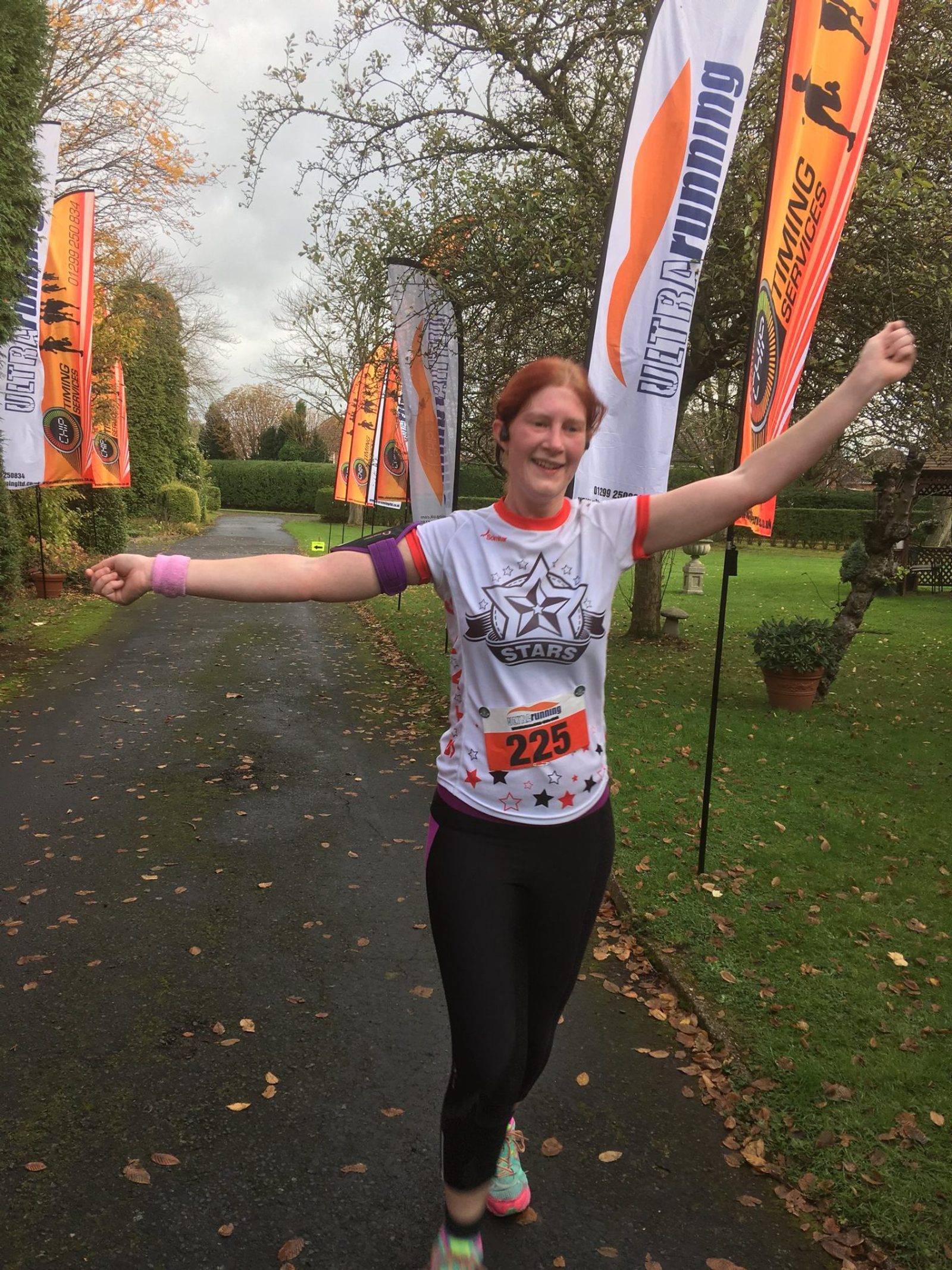 A person with red hair and a wide smile is seen running outdoors, arms raised in celebration. They are wearing a white "5 STAR$" shirt, black leggings, and sporting the number 235 on their chest. Bright orange and white race banners are visible in the background.