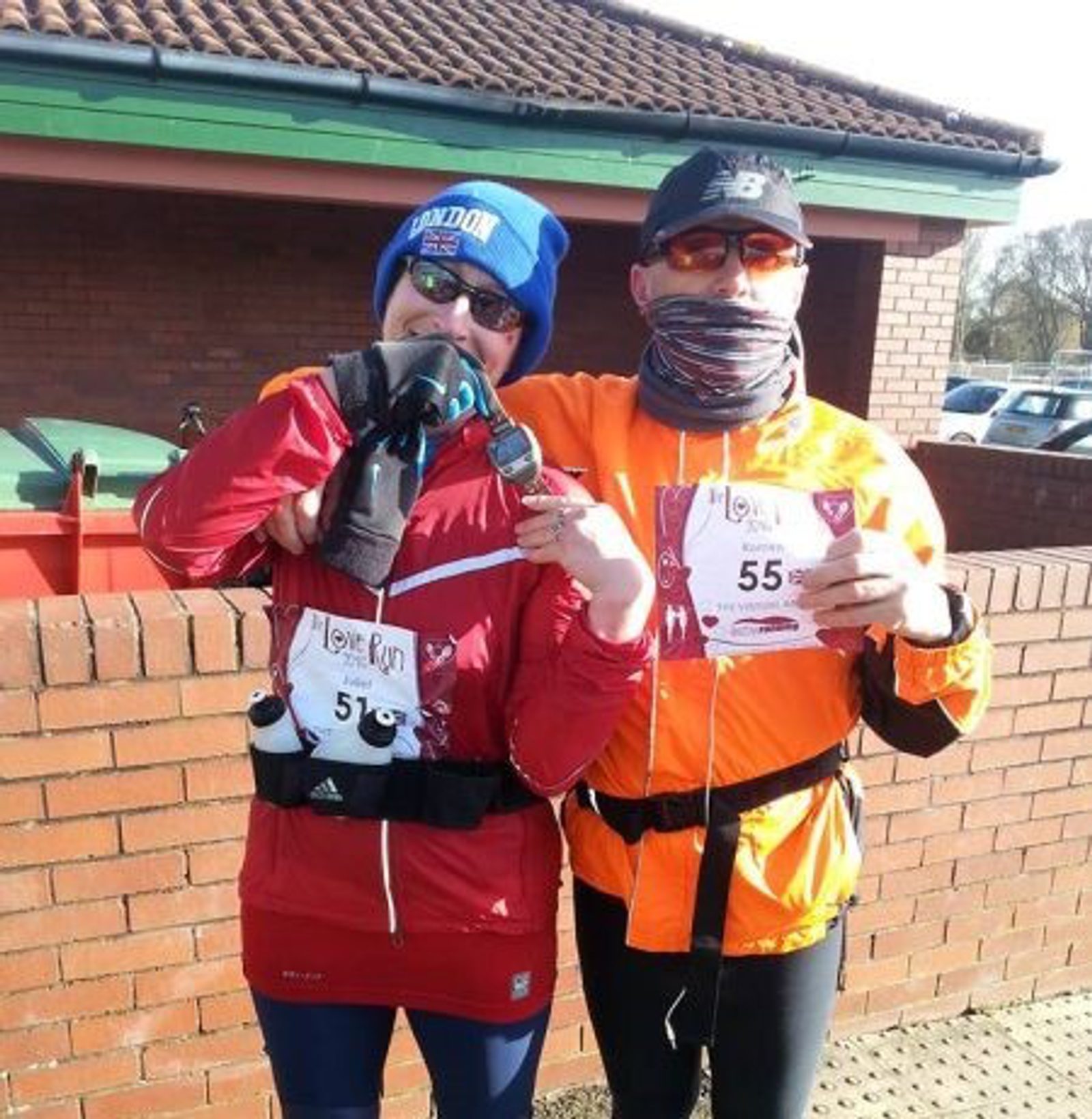 A runner in an orange jacket and a runner in a red jacket proudly display race bibs numbered 55 and 57, respectively. Both are dressed for cold weather, wearing hats, gloves, and sunglasses. They stand in front of a brick wall near a building with a brown roof.