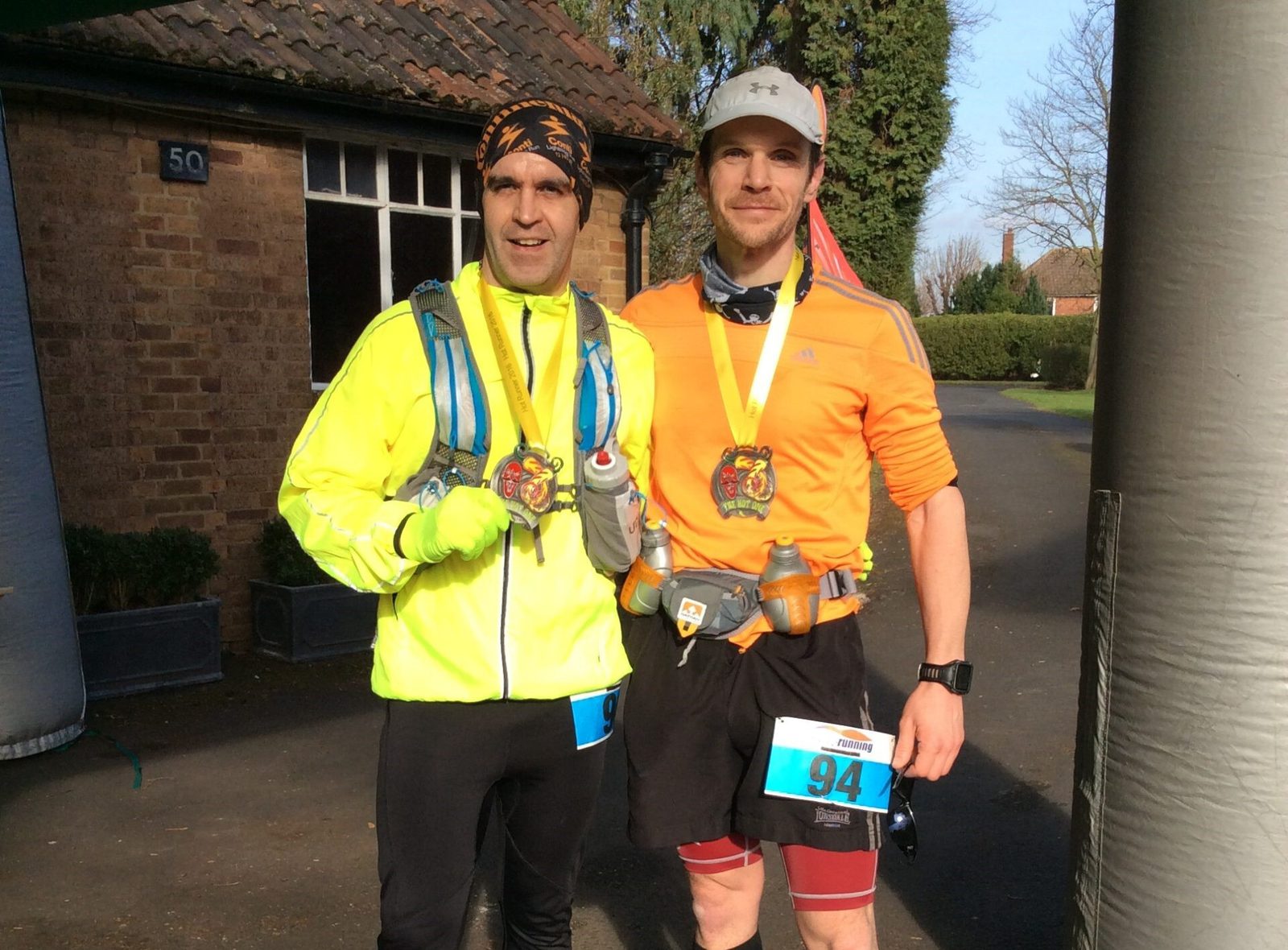 Two male runners, dressed in athletic gear, stand smiling with medals around their necks. The runner on the left wears a neon green jacket and black headband, while the one on the right sports an orange shirt and gray cap. Both hold their medals proudly in their hands.