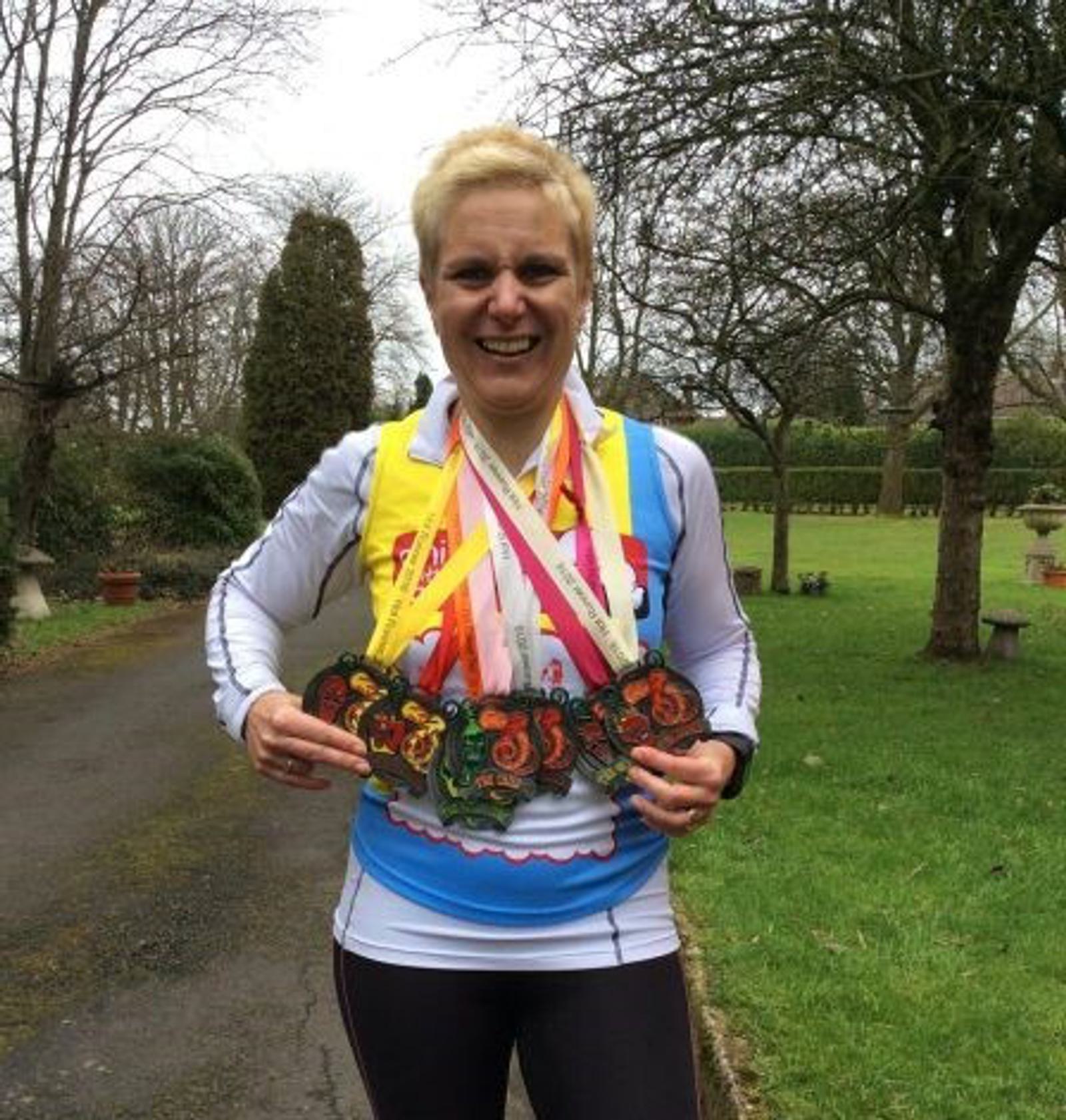 A person with short blonde hair is standing on a pathway in a park, smiling broadly. They are holding multiple medals around their neck, indicating participation in various events. The background features leafless trees and green grass.