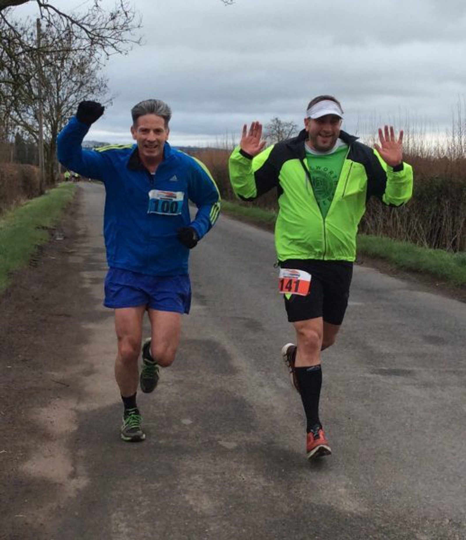 Two men, each wearing a race bib, run enthusiastically on a rural road. One man, in a blue jacket and shorts (bib number 100), has his left arm raised. The other man, in a neon yellow jacket and black shorts (bib number 141), appears to be waving with both hands up.