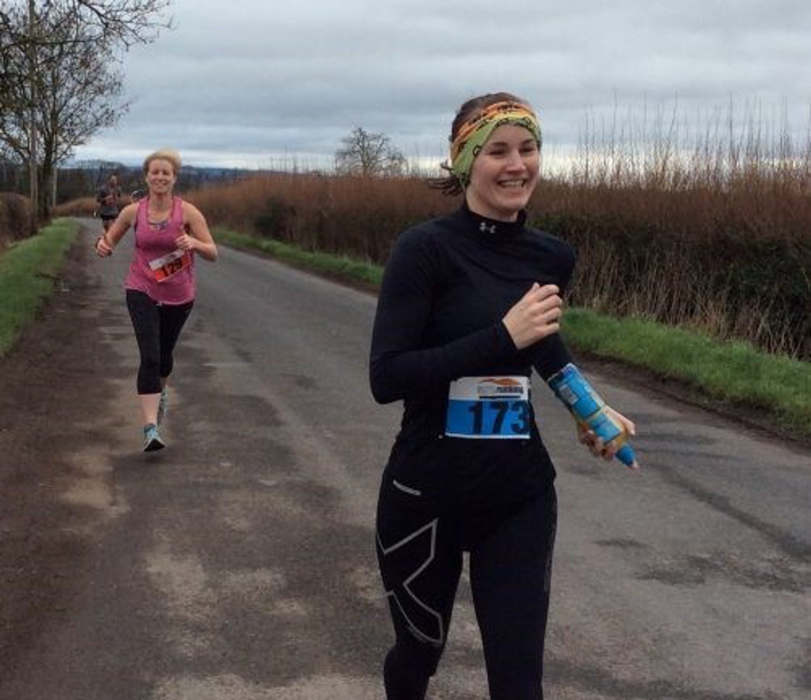 A person wearing a long-sleeve black top, dark leggings, and a colorful headband is running on a paved road holding a blue water bottle, smiling. Another runner in a pink top and black leggings is behind them. The sky is overcast and trees line the road.