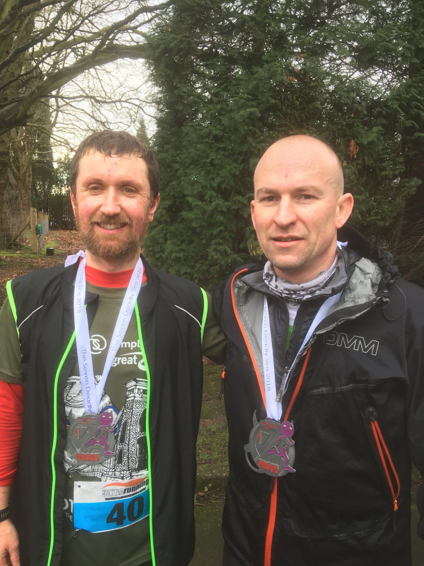 Two men, both wearing medals around their necks, stand outdoors after a race. The man on the left wears a green race bib labeled "40" while the man on the right wears a black jacket. They are smiling and appear to be in a wooded area.