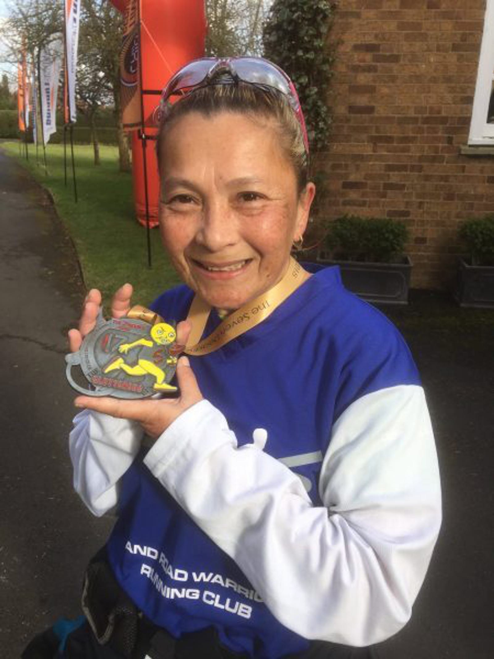A person with a headband and sunglasses on top of their head smiles while holding a race medal. They are wearing a blue and white long-sleeve shirt with "AND RAND WARFIH RUNNING CLUB" partially visible. Banners and a brick building are in the background.