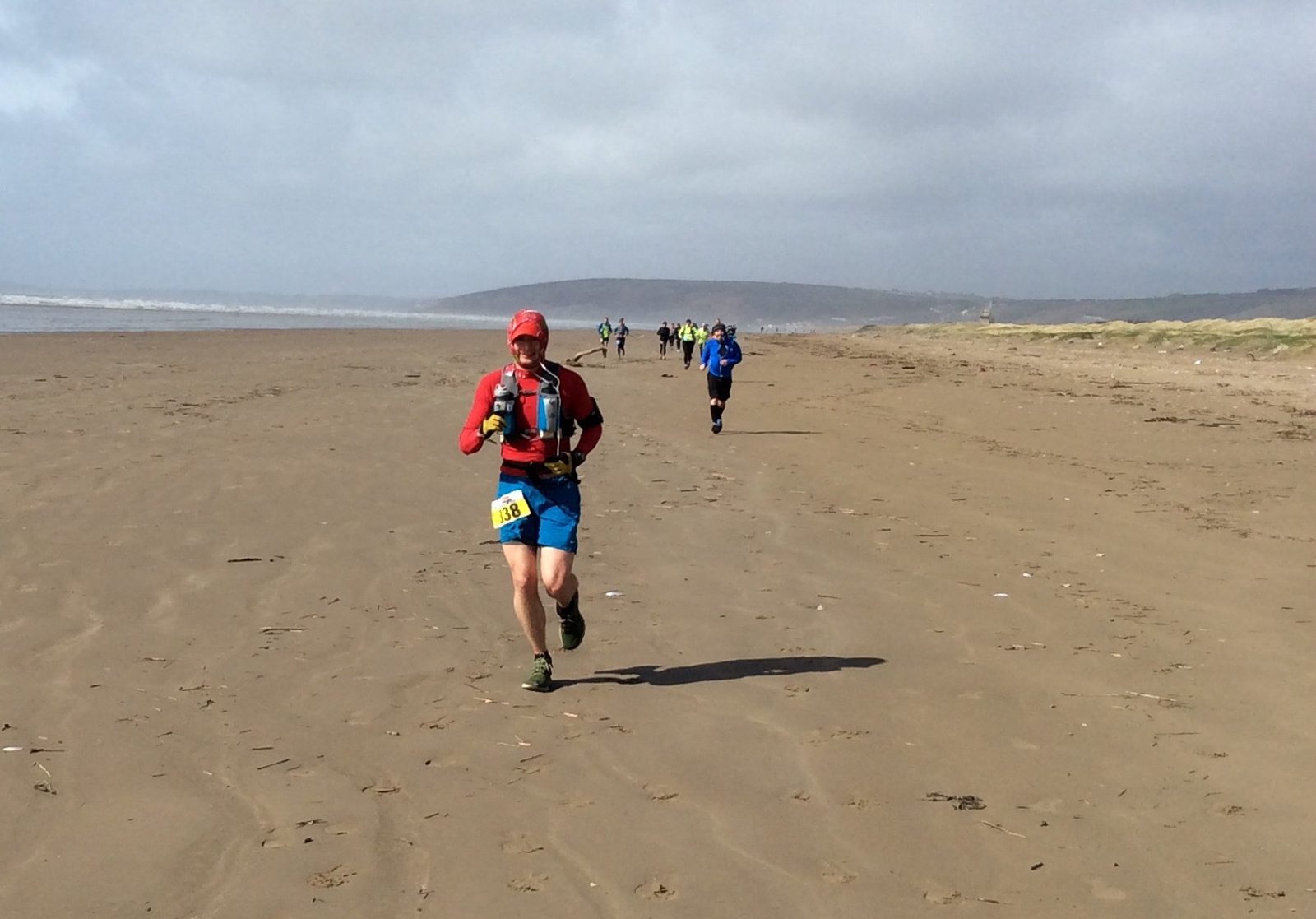A person wearing a red jacket, blue shorts, and a red hat runs on a sandy beach during a race. They have a number bib pinned to their shorts and a hydration pack on their back. Several other runners can be seen in the distance under a cloudy sky.