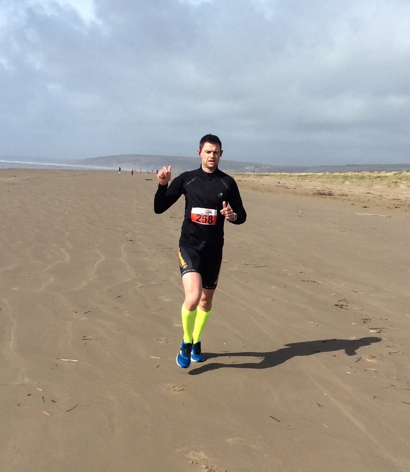A person running on a wide, empty beach under a cloudy sky. The runner is wearing bright yellow knee-high socks, blue shoes, black shorts, and a black long-sleeve top with a race number '258' attached to their waist.