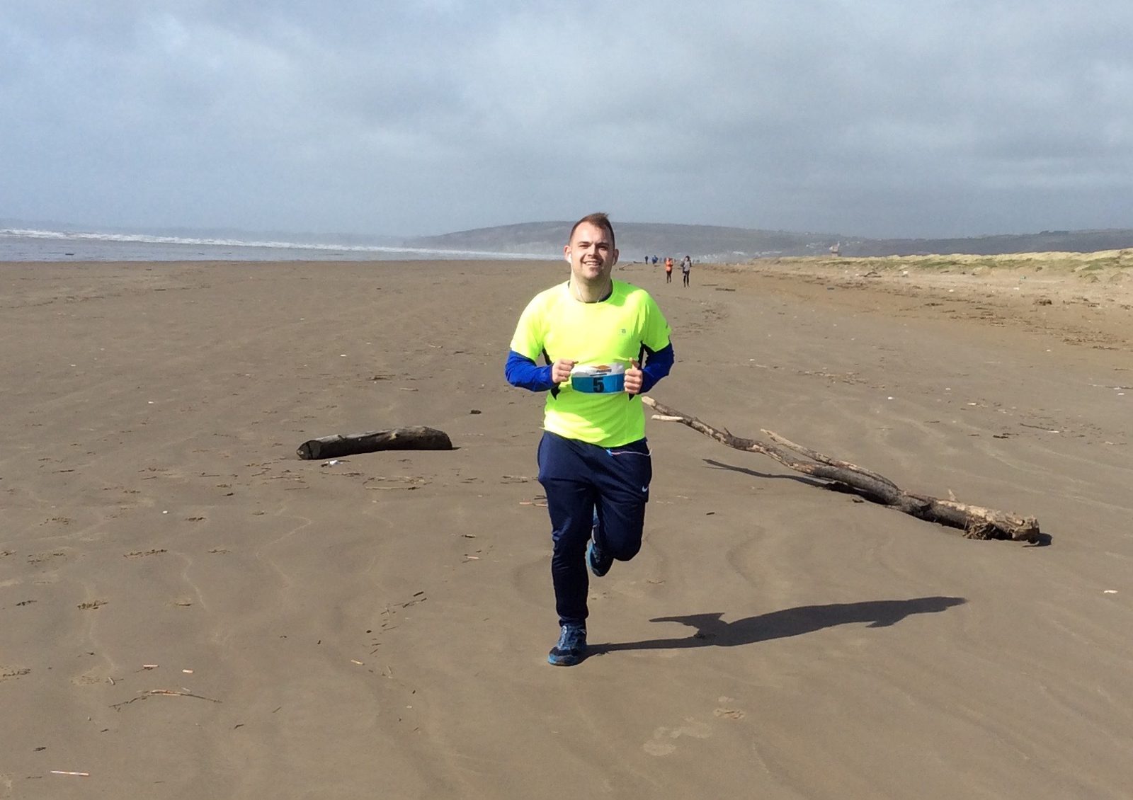 A person in a bright yellow shirt and blue pants is jogging on a wide, sandy beach. The sky is overcast, and there are a few pieces of driftwood scattered on the sand. In the background, there are a few other people on the beach and distant hills.
