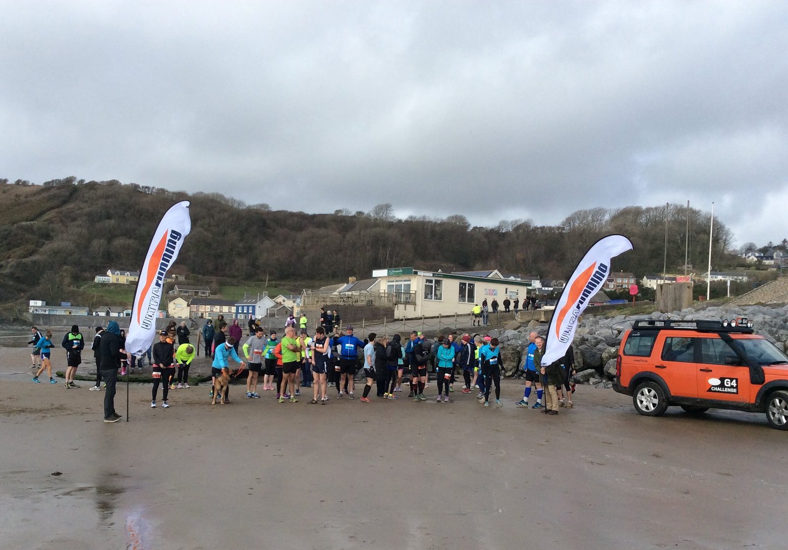 A group of runners gathers at the start line on a sandy beach, flanked by two white banners. Participants wear various colorful athletic clothing. A bright orange vehicle is parked nearby. In the background are buildings and a hillside with trees under a cloudy sky.
