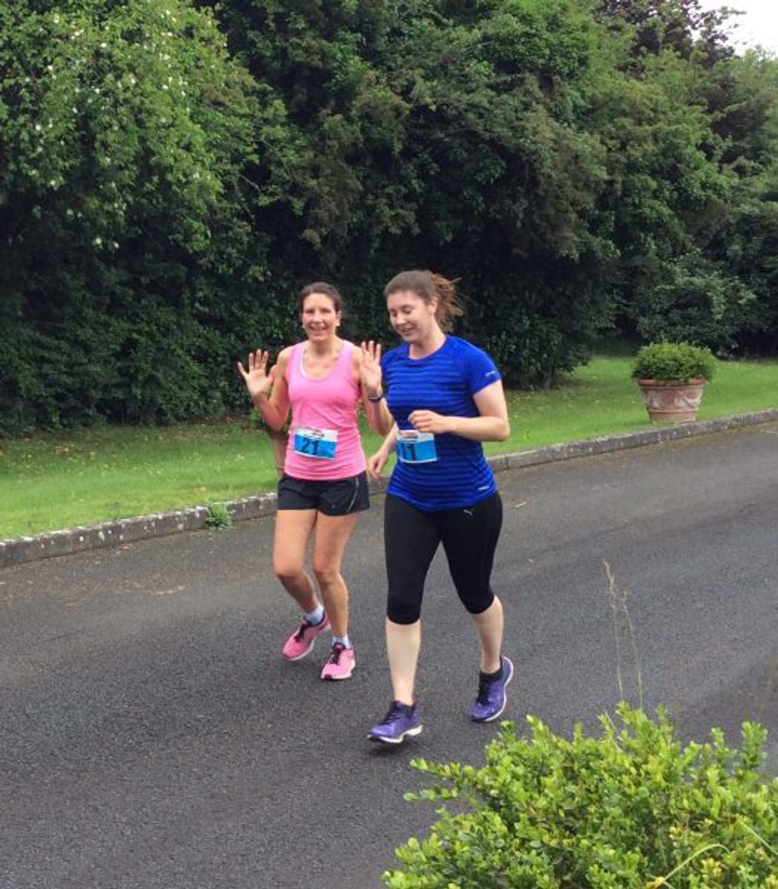 Two women are jogging on a paved path outdoors, surrounded by greenery. The woman on the left is wearing a pink shirt and shorts, while the woman on the right is in a blue shirt and black leggings. Both are smiling and waving at the camera.