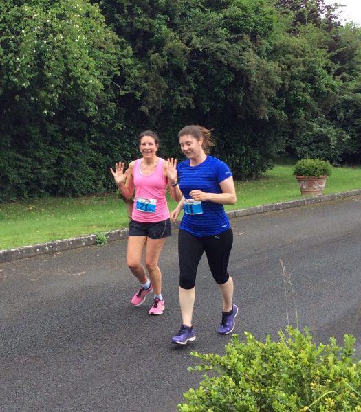 Two women are jogging on a paved path outdoors, surrounded by greenery. The woman on the left is wearing a pink shirt and shorts, while the woman on the right is in a blue shirt and black leggings. Both are smiling and waving at the camera.