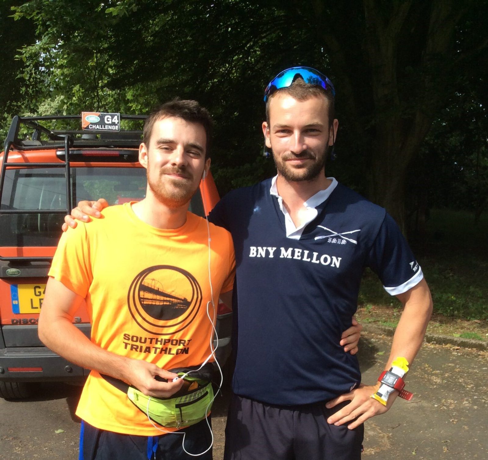 Two men pose for a photo outdoors. The man on the left is wearing an orange "Southport Triathlon" shirt and a fitness belt. The man on the right is wearing a navy blue shirt with "BNY MELLON" on it. They stand in front of a car with trees in the background.