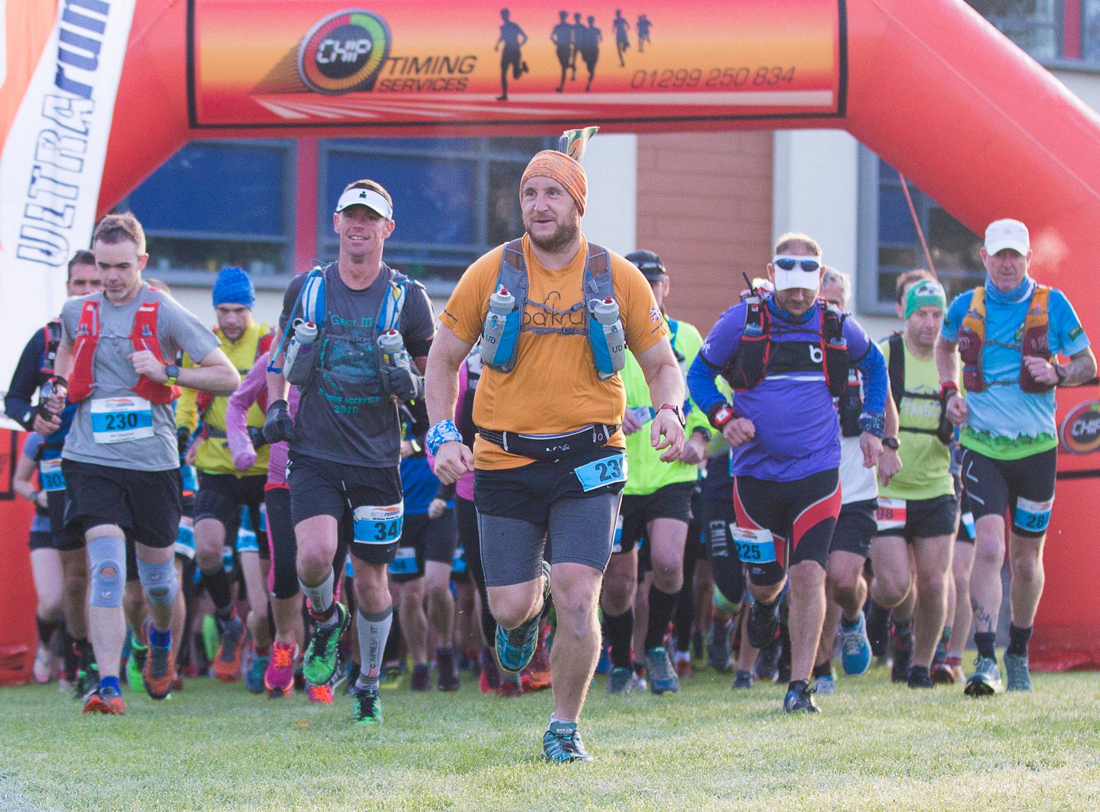 A group of runners, dressed in various gear, are participating in a race, captured in mid-stride. They are starting at an inflatable archway with banners overhead. The leading runner wears a yellow shirt and headband, while others follow closely behind.