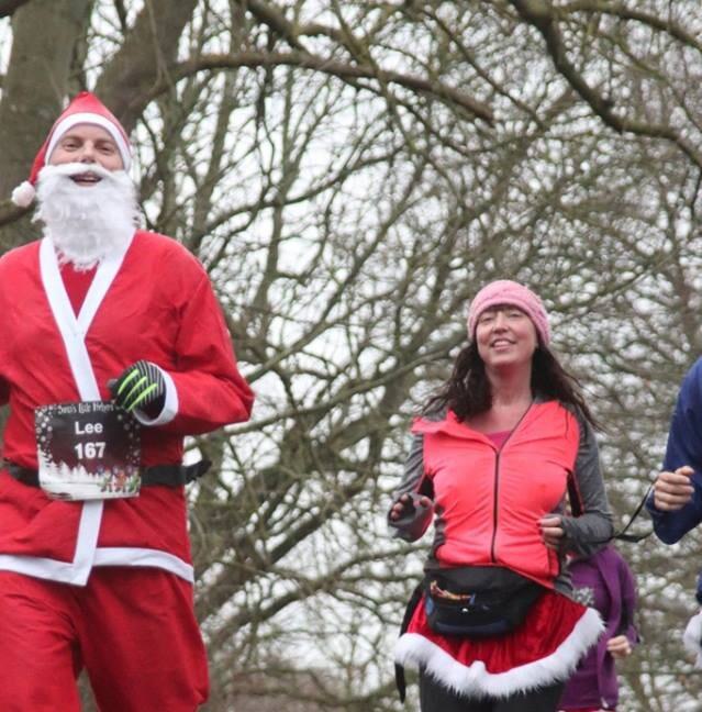 A participant dressed as Santa Claus and a woman in a pink hat and bright running gear jog together in a festive outdoor event. Trees without leaves are visible in the background, indicating a winter setting. Both appear to be enjoying themselves.