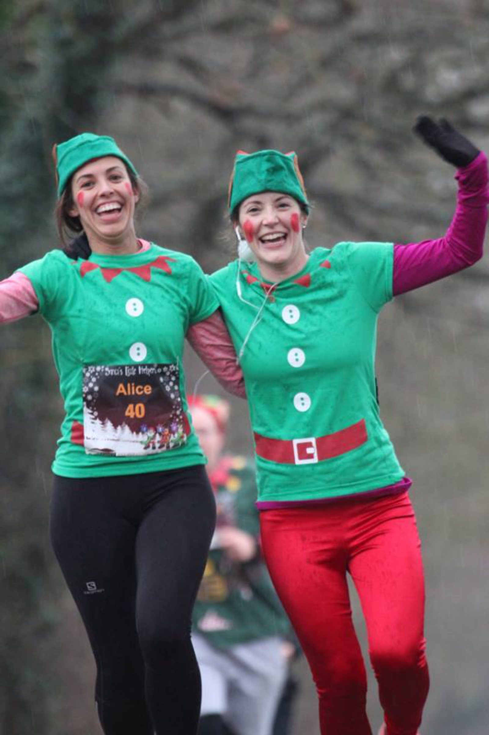 Two people dressed as Christmas elves are running outdoors. They are wearing green elf hats, green shirts with red and white trimmings, and elf belts printed on them. They are smiling, raising their arms, and appear to be participating in a fun holiday-themed race.
